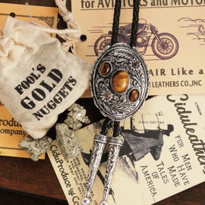 May include: An oval silver bolo tie with a tiger's eye stone center and three smaller stones. The tie has a black braided cord and decorative silver tips. A small bag of "Fool's Gold Nuggets" is next to the tie.