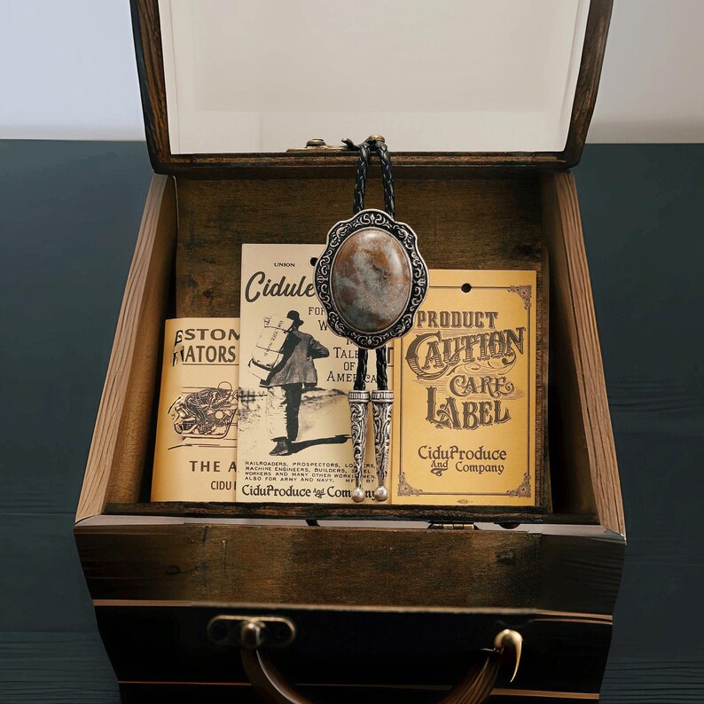 May include: A vintage-style bolo tie with an oval stone pendant and ornate silver-tone metalwork, displayed inside an open wooden box. The box contains aged paper ephemera with text and illustrations, creating a rustic, antique aesthetic.