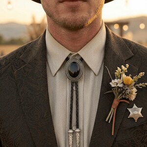 May include: A close-up of a person wearing a dark brown paisley patterned blazer, a light beige collared shirt, and a bolo tie with a black oval stone. A small bouquet and a sheriff's badge are pinned to the blazer.