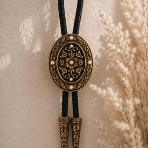 May include: An oval bolo tie with a black braided leather cord. The ornate pendant features a black background with a floral design in gold and small white pearls. The tips of the cord are gold with an engraved design.