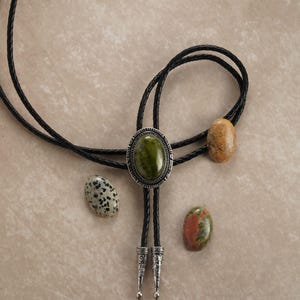 Puede incluir: Una corbata bolo con un gran colgante ovalado de piedra verde, rodeado por un marco de metal plateado. El cord&oacute;n de cuero negro se acompa&ntilde;a de tres piedras adicionales en tonos marr&oacute;n, beige y blanco moteado. La corbata presenta puntas decorativas de metal plateado.