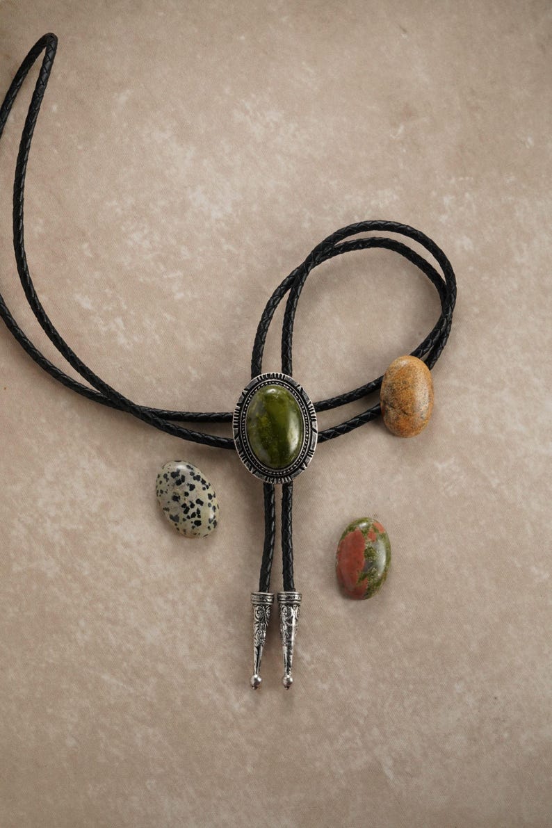 Puede incluir: Una corbata bolo con un gran colgante ovalado de piedra verde, rodeado por un marco de metal plateado. El cord&oacute;n de cuero negro se acompa&ntilde;a de tres piedras adicionales en tonos marr&oacute;n, beige y blanco moteado. La corbata presenta puntas decorativas de metal plateado.