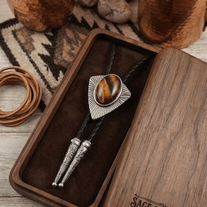 May include: A bolo tie with a tiger's eye stone set in a silver-colored triangle pendant, displayed in a wooden box. The box is open, revealing a dark brown interior. The box lid has the text "SAGE & SADDLE SIGNATURE".