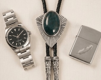 May include: A stainless steel wristwatch with a black dial, a silver lighter with a feather design, and a silver bolo tie with a dark green stone and black leather cord are displayed on a neutral background.