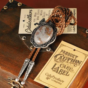 May include: A bolo tie with a silver-toned oval pendant featuring a marbled stone. The tie has a braided leather cord and decorative silver tips. Tags with text are visible.