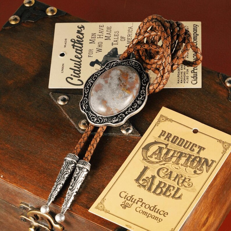 May include: A bolo tie with a silver-toned oval pendant featuring a marbled stone. The tie has a braided leather cord and decorative silver tips. Tags with text are visible.