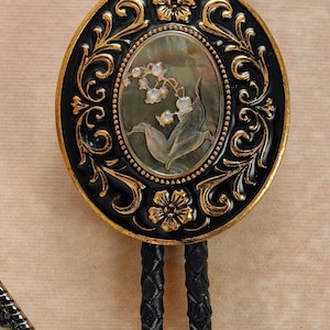 May include: Oval bolo tie pendant with a black and gold floral design. The center features a mother-of-pearl inlay with a lily of the valley illustration. The braided black leather cord has gold-colored metal tips. A decorative accessory.