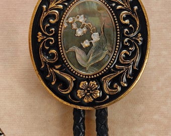 May include: Oval bolo tie pendant with a black and gold floral design. The center features a mother-of-pearl inlay with a lily of the valley illustration. The braided black leather cord has gold-colored metal tips. A decorative accessory.