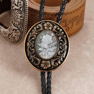May include: A black and gold bolo tie with a central oval design featuring a white rose carving. The oval is surrounded by ornate gold and black filigree. The tie has a braided black cord.