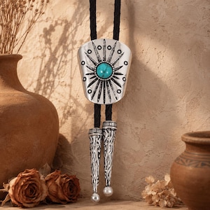 May include: A bolo tie featuring a silver-toned metal slide with a central turquoise stone and radiating sunburst engravings. The slide is attached to a black braided cord that terminates in two matching silver-toned metal tips with textured details.