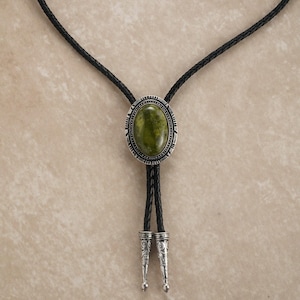 Puede incluir: Una corbata bolo con una piedra ovalada verde engastada en un marco de metal plateado. La corbata tiene un cord&oacute;n de cuero trenzado negro con puntas decorativas plateadas. El dise&ntilde;o es de estilo Western cl&aacute;sico.