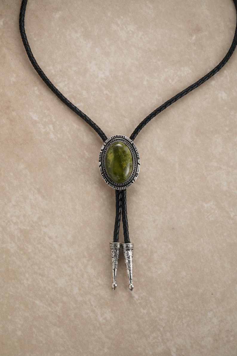 Puede incluir: Una corbata bolo con una piedra ovalada verde engastada en un marco de metal plateado. La corbata tiene un cord&oacute;n de cuero trenzado negro con puntas decorativas plateadas. El dise&ntilde;o es de estilo Western cl&aacute;sico.