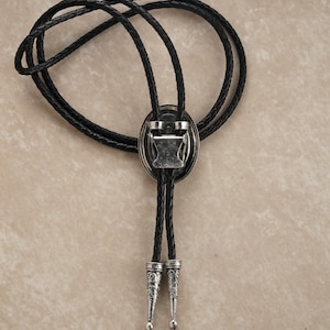 Puede incluir: Una corbata bolo de cuero negro con un cierre de metal plateado y puntas decorativas. El cierre tiene forma ovalada con un dise&ntilde;o detallado. Un accesorio Western cl&aacute;sico.