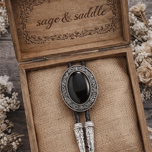 May include: A bolo tie with a large, oval black stone set in an ornate silver-tone frame. The tie is displayed in an open wooden box with the words "sage & saddle" engraved on the lid. The box is lined with burlap.