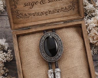 May include: A bolo tie with a large, oval black stone set in an ornate silver-tone frame. The tie is displayed in an open wooden box with the words "sage & saddle" engraved on the lid. The box is lined with burlap.