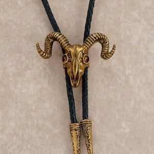 May include: A gold-colored ram skull bolo tie with a black braided leather cord. The ram skull has detailed horns and eye sockets. The cord ends are also gold-colored with an ornate design.