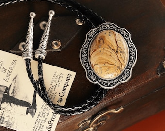 May include: A bolo tie with a braided black cord and silver tips. The focal point is an oval pendant with a patterned, tan-colored stone set in a decorative silver frame. A vintage-style tag is visible.