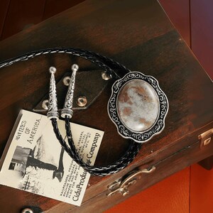 May include: A bolo tie with a braided black leather cord and silver-toned metal accents. The focal point is an oval pendant with a patterned stone set in a decorative frame. A vintage card with text is also visible.