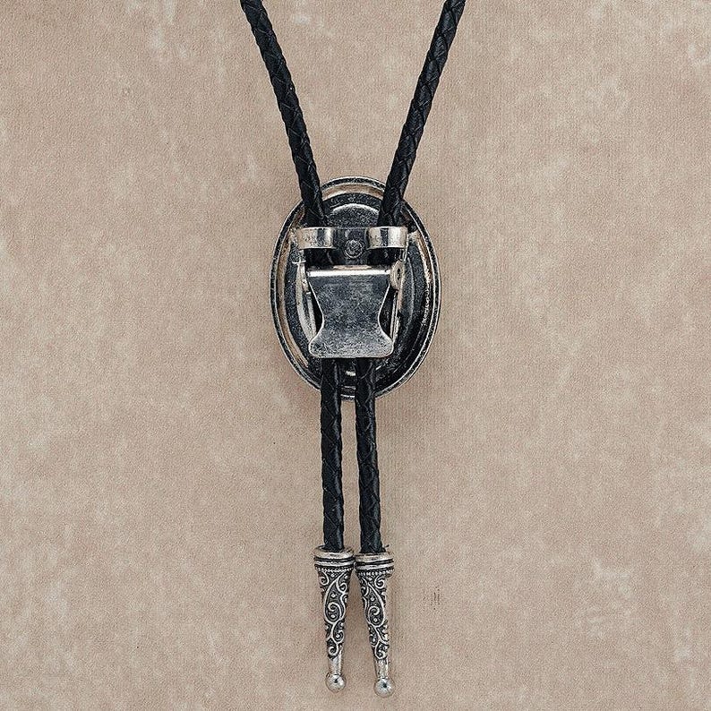 Puede incluir: Una corbata de lazo plateada con un cord&oacute;n de cuero trenzado negro. El colgante ovalado presenta un dise&ntilde;o ornamentado y un cierre. Las puntas del cord&oacute;n tambi&eacute;n son plateadas con un patr&oacute;n grabado. Un accesorio de estilo occidental.