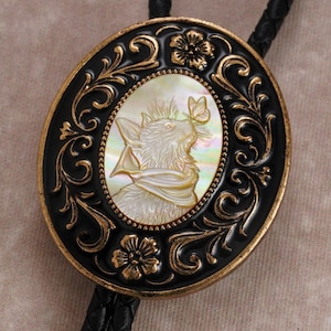 May include: Oval bolo tie with a black and gold filigree design. The center features a mother-of-pearl cameo of a wolf with a butterfly. The bolo tie has a black braided leather cord.