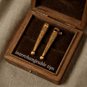 May include: Two gold-toned, conical metal tips featuring intricate engraved scrollwork patterns and rounded ends, presented inside a dark brown wooden box. The text "interchangeable tips" is printed in white on the dark velvet lining.