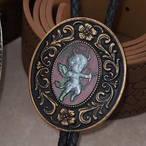 Puede incluir: Colgante de corbata bolo ovalado con un diseño de filigrana negro y dorado. El colgante presenta una imagen central de un querubín con arco y flecha. El querubín está en tonos grises y rosas, con un halo y pequeños cuernos. La corbata bolo tiene un cordón negro trenzado.