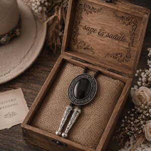 May include: A wooden box with the words "sage & saddle" engraved on the inside. The box contains a bolo tie with a black oval stone and silver-colored metal accents. The tie rests on a burlap-like fabric.