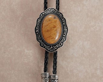 Puede incluir: Una corbata bolo con un colgante ovalado plateado y una piedra marrón claro. El colgante tiene un borde negro decorativo. El cordón trenzado negro tiene puntas plateadas. Accesorio de estilo occidental.