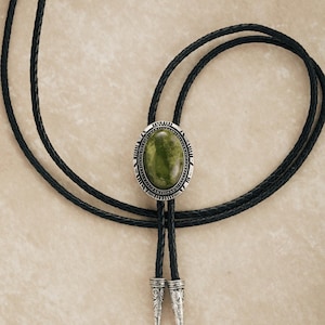Puede incluir: Una corbata bolo con una piedra ovalada verde engastada en un marco plateado. Tiene un cord&oacute;n de cuero trenzado negro y puntas decorativas plateadas. El dise&ntilde;o recuerda a la vestimenta del oeste.