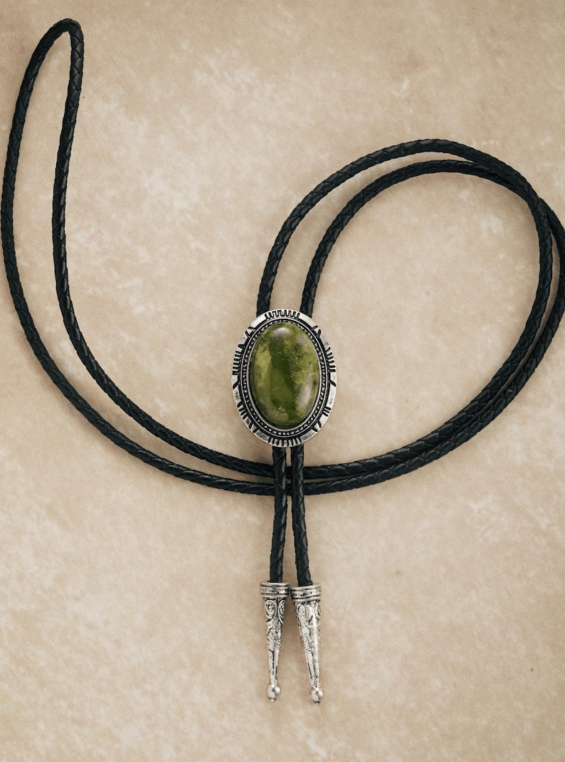 Puede incluir: Una corbata bolo con una piedra ovalada verde engastada en un marco plateado. Tiene un cord&oacute;n de cuero trenzado negro y puntas decorativas plateadas. El dise&ntilde;o recuerda a la vestimenta del oeste.