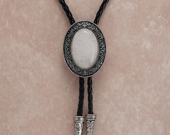 May include: A bolo tie with a white oval stone set in a decorative silver-tone frame. The black braided cord has silver-tone tips. The bolo tie is a Western-style accessory.