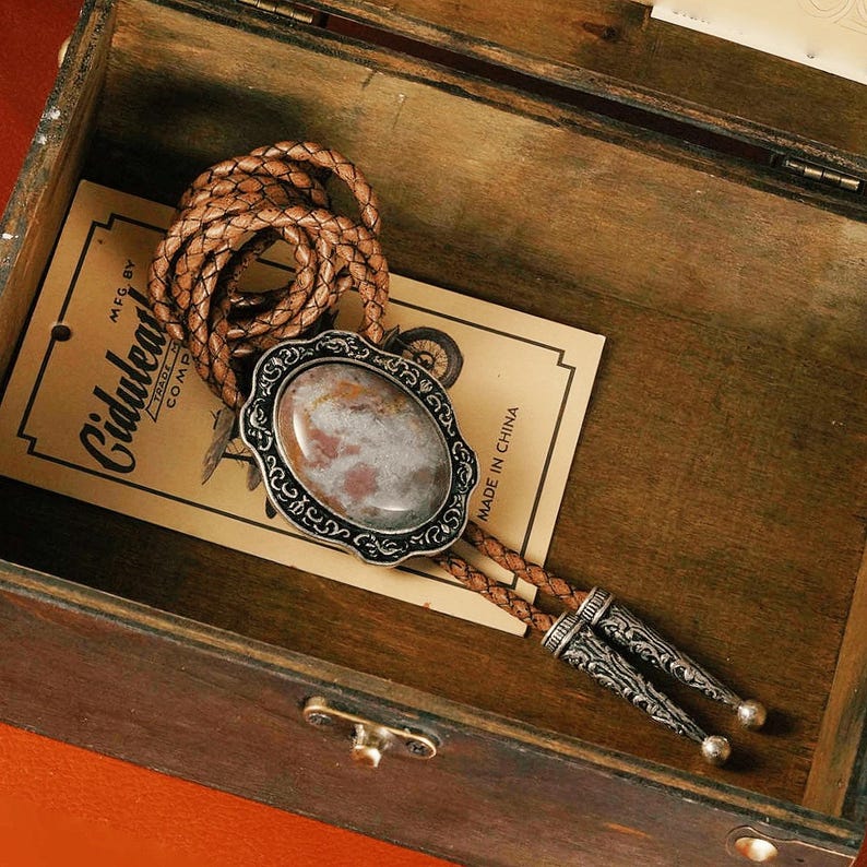 May include: A bolo tie with a brown braided leather cord and a decorative silver-toned slide. The slide features an oval stone with brown and gray hues, set in a detailed silver frame. The tie is displayed inside a wooden box.