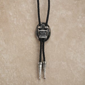 Puede incluir: Una corbata bolo con un cord&oacute;n de cuero trenzado negro y un colgante ovalado plateado. El colgante tiene un dise&ntilde;o decorativo y un cierre para ajustar la longitud del cord&oacute;n. Las puntas del cord&oacute;n tambi&eacute;n son plateadas.