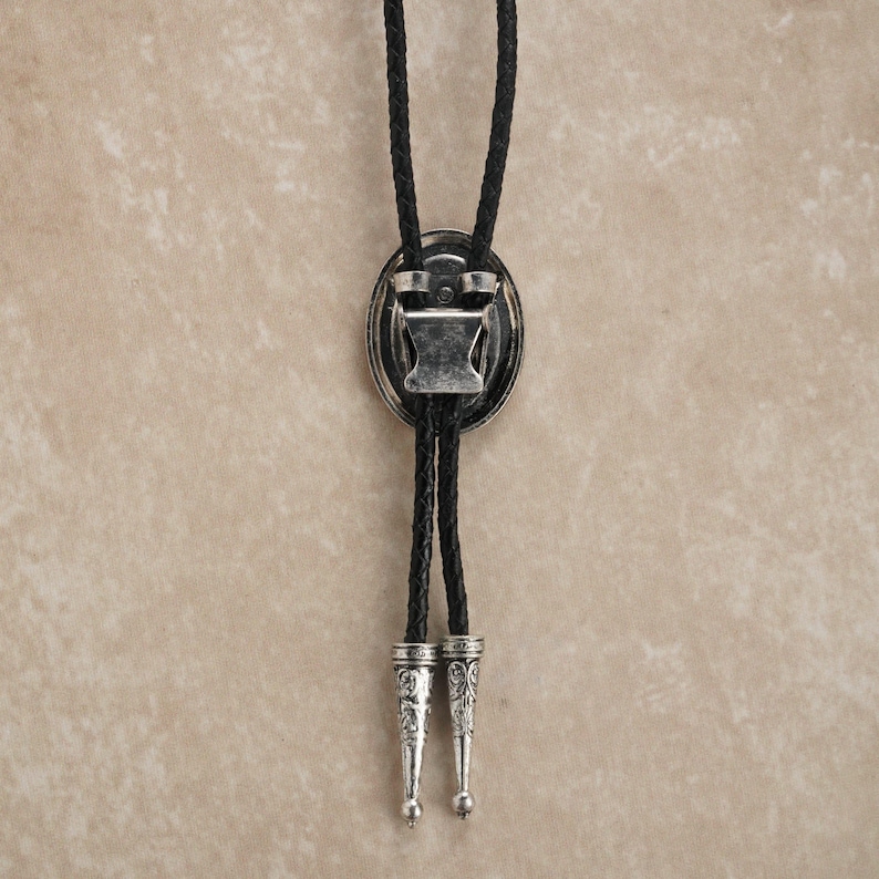 Puede incluir: Una corbata bolo con un cord&oacute;n de cuero trenzado negro y un colgante ovalado plateado. El colgante tiene un dise&ntilde;o decorativo y un cierre para ajustar la longitud del cord&oacute;n. Las puntas del cord&oacute;n tambi&eacute;n son plateadas.