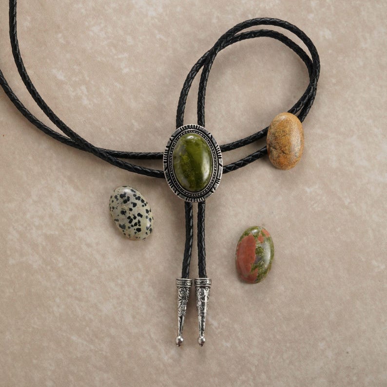 Puede incluir: Una corbata bolo de cuero negro con un colgante ovalado plateado con una piedra verde. La corbata va acompa&ntilde;ada de tres piedras ovaladas en tonos beige, verde y blanco moteado. La corbata bolo tiene puntas plateadas decorativas.
