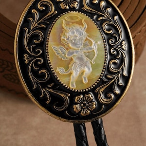 May include: Oval bolo tie with a black and gold frame. The center features a carved cameo of a cherub with a bow and arrow. The cherub is surrounded by a halo. The tie has a black braided cord.