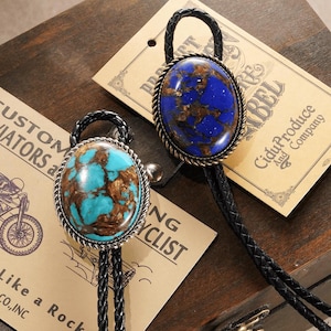 Peut inclure: Deux cravates bolo avec des pierres ovales. L'une présente une pierre turquoise veinée de brun, l'autre une pierre bleue avec des inclusions brunes. Les deux ont des bordures argentées en forme de corde et des cordons en cuir tressé noir. Des étiquettes vintage sont en arrière-plan.