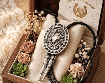 May include: A Western-themed wedding gift set in a wooden box. The set includes a silver bolo tie with an oval pendant and a black braided leather cord. The box also contains dried flowers, succulents, and a card that reads "Western Wedding".