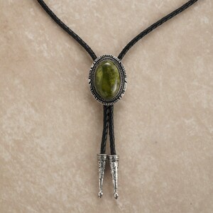 Puede incluir: Una corbata bolo con una piedra ovalada verde engastada en un marco ovalado plateado. El cord&oacute;n de cuero trenzado negro tiene puntas decorativas plateadas. La corbata se muestra sobre un fondo neutro.