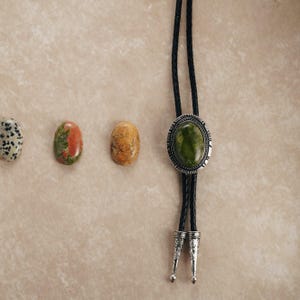 Puede incluir: Una corbata bolo con un colgante de piedra ovalada verde y un cord&oacute;n trenzado negro. El colgante est&aacute; enmarcado por metal plateado. Tres piedras preciosas ovaladas en tonos marr&oacute;n, rojo y blanco est&aacute;n dispuestas a la izquierda.