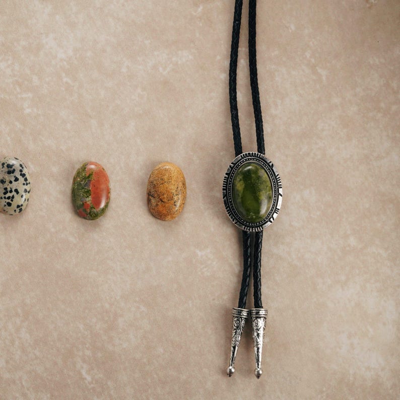 Puede incluir: Una corbata bolo con un colgante de piedra ovalada verde y un cord&oacute;n trenzado negro. El colgante est&aacute; enmarcado por metal plateado. Tres piedras preciosas ovaladas en tonos marr&oacute;n, rojo y blanco est&aacute;n dispuestas a la izquierda.