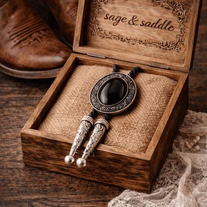 May include: A bolo tie with a black oval stone and silver-toned metal accents rests in a wooden box lined with burlap. The box lid is engraved with the words "sage & saddle". Brown leather cowboy boots are visible in the background.