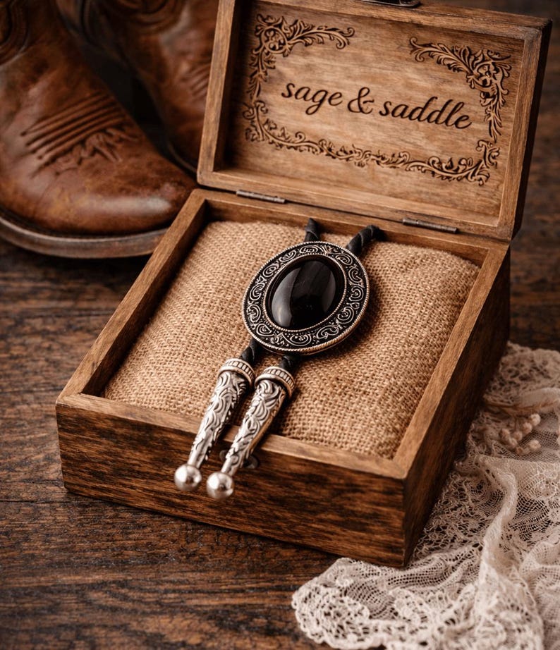 May include: A bolo tie with a black oval stone and silver-toned metal accents rests in a wooden box lined with burlap. The box lid is engraved with the words "sage & saddle". Brown leather cowboy boots are visible in the background.