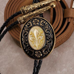 Puede incluir: Una corbata bolo negra y dorada con un colgante ovalado con un diseño de flor de lis. El colgante está rodeado de adornos ornamentales y sobre un fondo crema. La corbata tiene un cordón negro trenzado y puntas doradas.