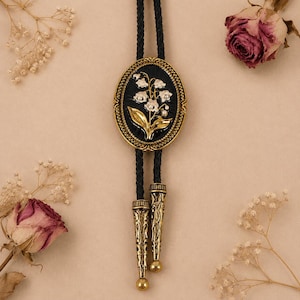 May include: A bolo tie featuring an oval black centerpiece with a gold-toned lily of the valley floral motif and small white accents. The centerpiece is framed in a gold-toned metal border and attached to a black braided cord with two long, ornate gold-toned metal tips at the ends.