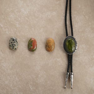 Puede incluir: Una selecci&oacute;n de piedras preciosas pulidas y una corbata bolo. Las piedras preciosas incluyen una piedra blanca y negra moteada, una piedra roja y verde y una piedra de color canela. La corbata bolo tiene una piedra ovalada verde engastada en un metal plateado.
