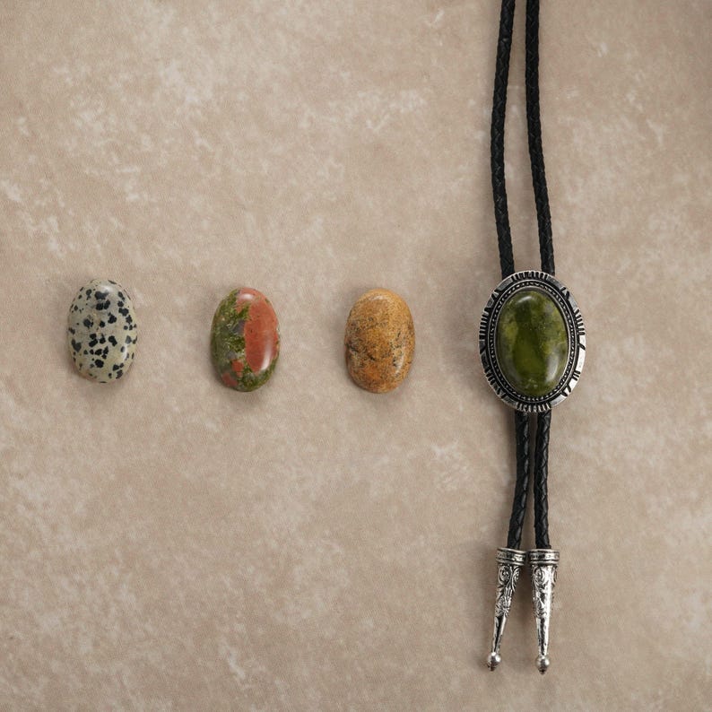 Puede incluir: Una selecci&oacute;n de piedras preciosas pulidas y una corbata bolo. Las piedras preciosas incluyen una piedra blanca y negra moteada, una piedra roja y verde y una piedra de color canela. La corbata bolo tiene una piedra ovalada verde engastada en un metal plateado.