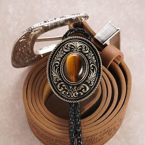 Puede incluir: Un cinturón de cuero marrón con una hebilla plateada y una corbata bolo decorativa. La corbata bolo presenta un colgante circular, ornamentado, de color bronce con una piedra ojo de tigre. El cordón negro trenzado cuelga sobre el cinturón.