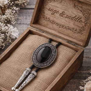 May include: A bolo tie with a large, oval black stone set in an ornate silver-colored frame, displayed in an open wooden box lined with burlap. The box is engraved with the words "sage & saddle".