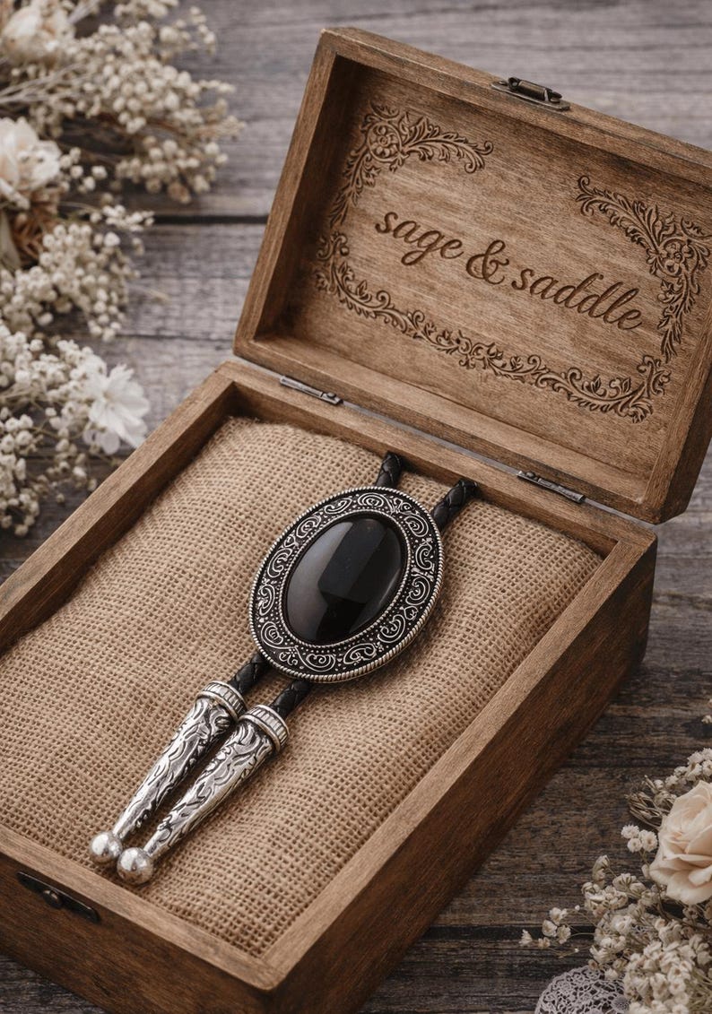 May include: A bolo tie with a large, oval black stone set in an ornate silver-colored frame, displayed in an open wooden box lined with burlap. The box is engraved with the words "sage & saddle".